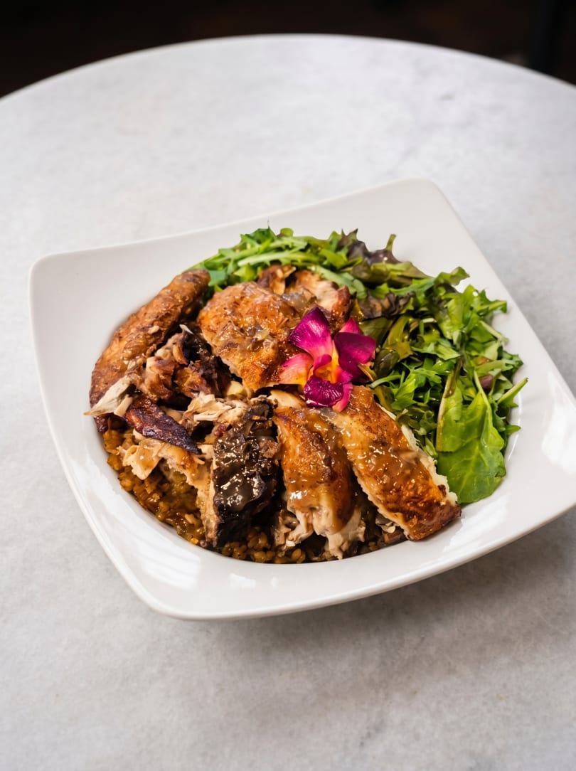 A white square plate holds roasted chicken, rice, mixed greens, and a purple orchid garnish.