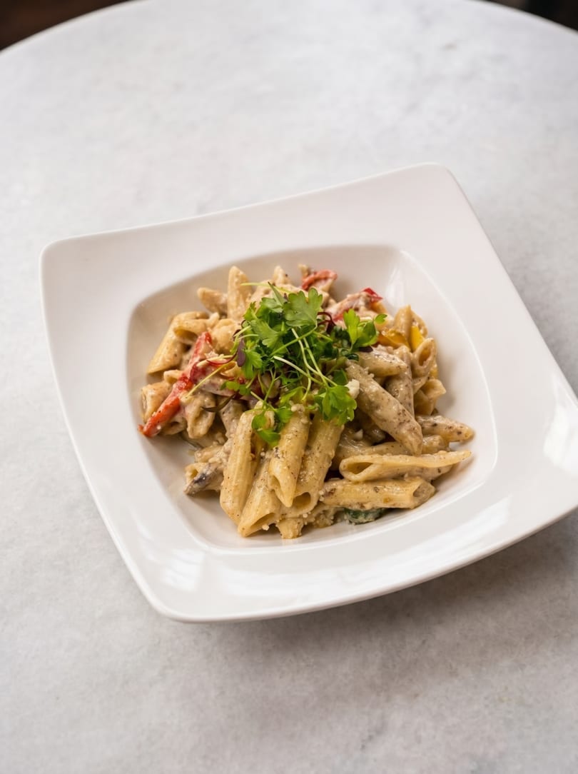 Creamy penne pasta with red peppers and fresh herbs in a white square bowl on a light table.