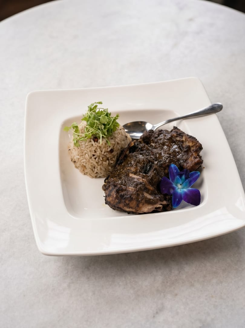 Jerk chicken with rice and peas, garnished with cilantro and a blue orchid, on a white plate.